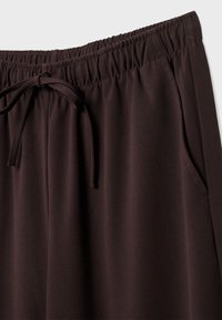 Dunkelbraune Hose mit elastischem Bund, Schnürung, Seitentaschen und glatter Textur. Nahaufnahme des Bundes und des Stofffalls.