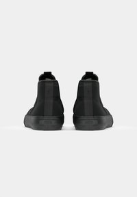 Zapatillas altas negras con una textura suave, suela de goma plana y marca en el talón. Diseño minimalista con costuras verticales sutiles.