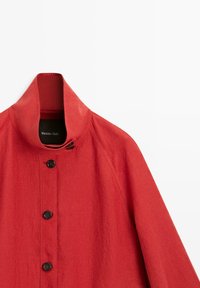 Rode jas met hoge kraag, zwarte knopen aan de voorkant en een knoopsluiting bij de hals.