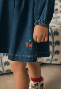 Denimkjole med puf-ærmer, der har rød kirsebærsbroderi på sømmen og kontrastfarvet guldstikning. Foldet manchet detalje synlig.