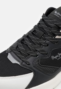 Chaussure de sport noire en mesh et cuir, avec accents texturés, détails gris et lacets à pois. Logo Pepe Jeans sur le côté.