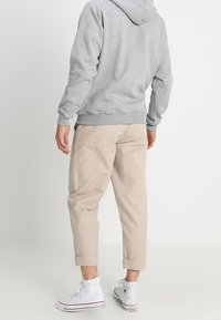Homme debout, de dos, portant un sweat à capuche gris, un pantalon beige retroussé et des baskets blanches montantes Converse sur un sol blanc.