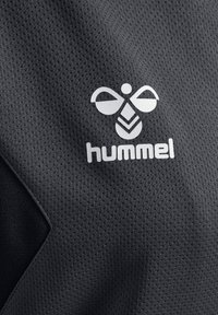 Zbliżenie na czarną tkaninę sportową z białym logo hummel, z stylizowaną pszczołą nad nazwą marki.