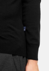 Pull en tricot noir avec une coupe ajustée, des poignets côtelés et une petite étiquette bleue sur le côté. Texture douce et silhouette classique.