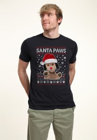 Svart t-shirt med en grafik av en hund i en röd tomteluva med texten "SANTA PAWS", omgiven av snöflingor och tassavtryck.