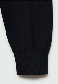 Gros plan sur la manche d'un pull noir en maille avec un poignet côtelé sur un fond clair uni.