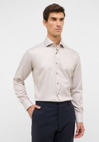 Camicia beige chiaro a maniche lunghe con un motivo sottile, colletto e bottoni marroni, abbinata a pantaloni scuri.