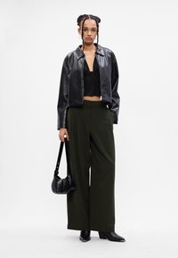 Γυναίκα με πλεγμένα μαλλιά, φορεμένη μαύρη δερμάτινη μπουφάν, μαύρη κοντή μπλούζα, φαρδιά σκούρα πράσινα παντελόνια, μαύρες μπότες και κρατώντας μια μικρή μαύρη τσάντα.