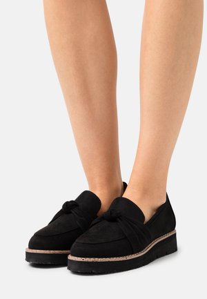 Schwarze Wildleder-Loafer mit einem geknoteten Detail, ausgestattet mit einer dicken, strukturierten Sohle und einem subtilen hellen Akzent entlang der Unterseite.