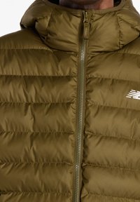 Homme portant une veste matelassée vert olive avec capuche et fermeture éclair frontale, montrant un logo partiel sur la poitrine.