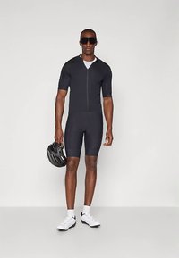 Czarny strój kolarski z krótkimi rękawami, zamek błyskawiczny z przodu i obcisłe spodenki. Noszony z kaskiem i białymi butami kolarskimi. Gładki, odprowadzający wilgoć materiał.