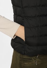 Czarna kamizelka puchowa z pikowanym wzorem i bocznymi kieszeniami, nałożona na jasnoszary, długi rękaw, w połączeniu z teksturowanymi oliwkowymi spodniami.