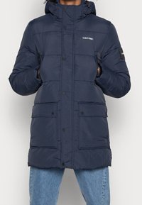 Granatowa pikowana kurtka z kapturem, zapinana na zamek i zatrzaski, z dwoma przednimi kieszeniami oraz naszywką z logo na rękawie. Gładka tekstura.