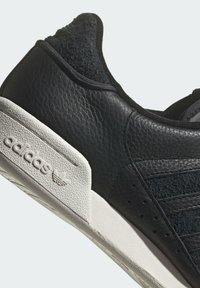 Černá kožená teniska s texturovaným povrchem, vybavená bílou gumovou podešví, designem tří pruhů a diskrétním logem na patě.