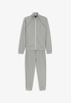 Svetlo siva dvodelna trenirka z zadrgo na jakni in hlačami z dodatnim trakom za vezanje, s tankimi črnimi obrobami in stranskimi žepi.
