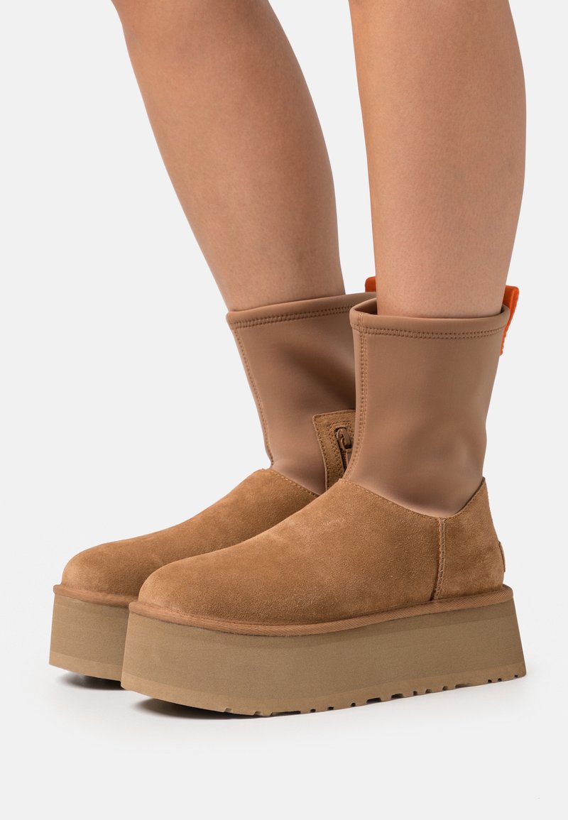 Paire de bottes plateformes couleur beige avec la partie du pied en daim et une tige supérieure lisse, portées aux jambes d'une personne sur un fond uni.