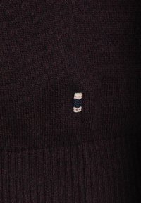 Jersey de punto en burdeos profundo, con textura acanalada. Etiqueta pequeña con una banda navy y acentos rojos visible en el costado.