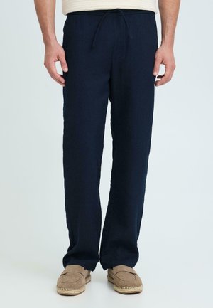 Hombre con pantalones azul marino con cordón y mocasines beige, de pie frente a un fondo claro y liso.