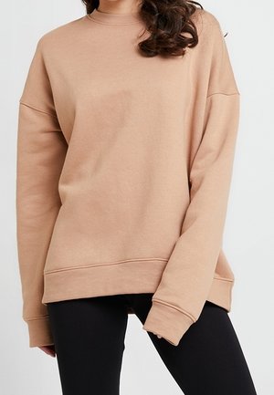 Kobieta ubrana w luźną, beżową bluzę z długim rękawem i czarne legginsy, stojąca na tle jednolitego białego tła.