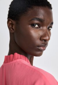 Rosa Bluse mit plissiertem Ausschnitt, glatter Textur und rundem Kragen. Der Hautton des Models ist dunkel und die Haare sind kurz und lockig.