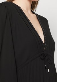 Abito nero con profondo scollo a V, texture plissettata, maniche arricciate e dettagli in metallo lungo il décolleté. Accento in vita con nodo.