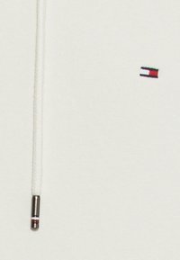 Felpa bianca con cappuccio dotata di cordino con fermaglio metallico. Sul lato sinistro del petto è presente un piccolo logo multicolore in rettangoli rossi, bianchi e verdi.