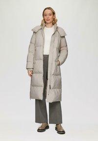 Pufferjas in lichtbeige met een rits aan de voorkant en een oversized kraag. Heeft een gewatteerd ontwerp en twee zijzakken. Gestyled over een grijze broek.
