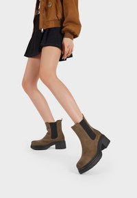 Stivaletti in suede marrone con lati elastici neri e un tab per il tiraggio, caratterizzati da una suola spessa nera e una texture liscia. Indossati con una gonna di jeans.