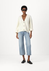 Kreminė kabelinė megztinė su giliu V formos iškirpimu, dideliais mygtukais ir sukniubuotais rankogaliais, derinama su šviesiai mėlynomis plačiomis kelnėmis ir juodais loafer'iais.