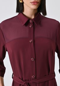 Robe chemise bordeaux avec col, manches trois-quarts et taille nouée. Présente des boutons ronds et une texture de tissu lisse.