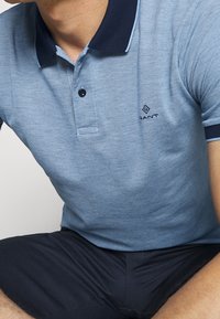 Polo bleu clair en tissu texturé, doté d'un col bleu foncé, de trois boutons et d'un logo sur le côté gauche de la poitrine.