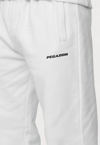 Bílé tepláky vyrobené z bavlněné směsi, s boční kapsou a černým potiskem loga "PEGADOR" na stehně. Měkká textura a volný střih.