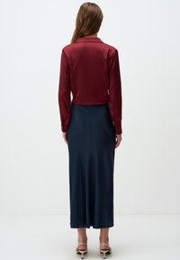 Camicia di raso bordeaux con colletto, abbinata a una lunga gonna di raso blu navy. Modella che indossa tacchi chiari, evidenziando una texture liscia.