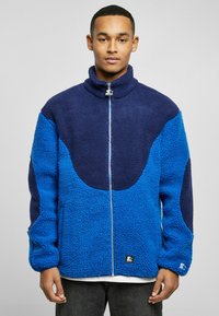 Fleece jacket featuring a zip front, high collar, and a color-block design with shades of blue and navy. Textured with a plush finish.