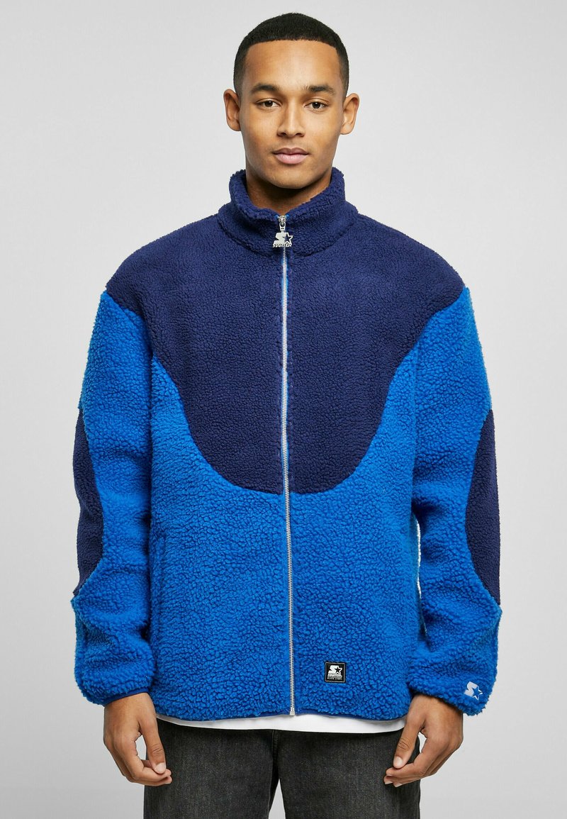 Fleece jacket featuring a zip front, high collar, and a color-block design with shades of blue and navy. Textured with a plush finish.