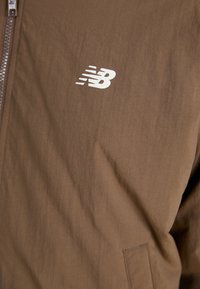 Casaco leve castanho com uma textura suave, apresentando um logótipo branco da New Balance no lado esquerdo do peito. Fecho com zíper e design minimalista.