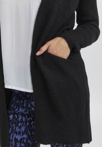 Črn cardigan iz mehkega pletenega materiala z dolgimi rokavi, odprtim sprednjim delom in stranskimi žepi. Pod njim je delno viden bel bluzon.