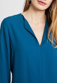 Bluzka w kolorze morskim z dekoltem w kształcie litery V i długimi rękawami, wykonana z gładkiej tkaniny. Charakteryzuje się delikatnym zagięciem przy dekolcie i minimalistycznym designem.