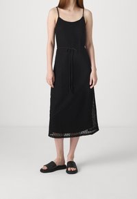 Černé pletené šaty s tenkými ramínky, délky do poloviny lýtek a s páskem na stahování v pase. Doplněné jednoduchými černými sandály na klínu.