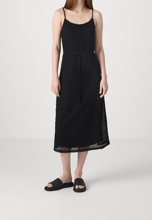 Zwarte gebreide jurk met dunne bandjes, tot halverwege de kuit lengte en een koord in de taille. Gecombineerd met eenvoudige zwarte instapsandalen.