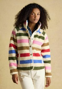 Žena oblečená v farebnej pruhovanej fleecovej bunde s patentkami, preoblečená cez denimovú košeľu a krémové nohavice, stojaca na jednoliatom pozadí.