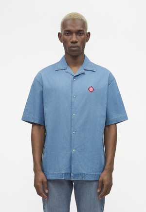 Νεαρός άνδρας με κοντά ξανθά μαλλιά που φοράει ένα μπλε κοντομάνικο πουκάμισο με κουμπιά και ένα κόκκινο λογότυπο στο αριστερό στήθος.