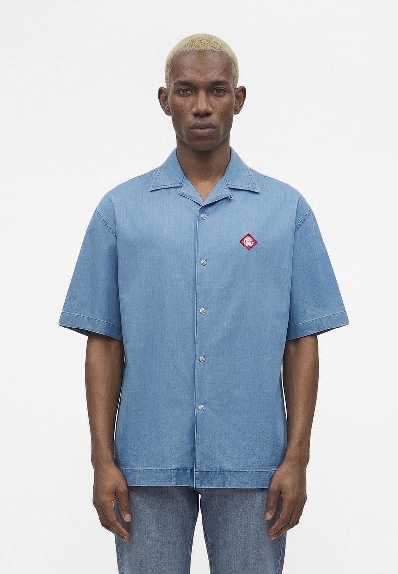 Νεαρός άνδρας με κοντά ξανθά μαλλιά που φοράει ένα μπλε κοντομάνικο πουκάμισο με κουμπιά και ένα κόκκινο λογότυπο στο αριστερό στήθος.