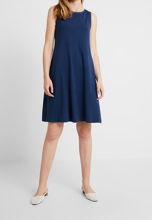 Femme debout portant une robe sans manches bleu marine au genou et des chaussures blanches tissées à talons bas, sans lacets, sur un fond uni.