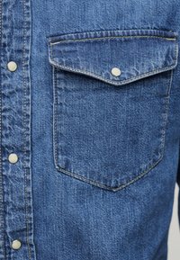 Τσέπη μπλε τζιν πουκάμισου με καπάκι και μεταλλικό κουμπί με πατητό κούμπωμα, κοντινή εικόνα που δείχνει την υφή του υφάσματος και μέρος της κουμπωμένης μπροστινής ταινίας.