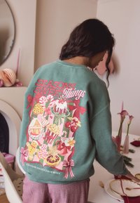Žalias džemperis su spalvotu grafiniu elementu, kuriame yra užrašas „SEASONS SWEET Things“ ir dekoratyviniai šventiniai saldumynai. Turi laisvą kirpimą ir apvalų kaklo iškirpimą.