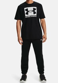 Hombre vistiendo camiseta negra de Under Armour con logo y texto, pantalones deportivos negros con logo y zapatillas negras sobre fondo blanco.