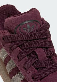 Gros plan d'une basket en daim bordeaux avec des lacets bordeaux texturés, des rayures grises et le logo trefoil noir d'Adidas sur la languette.