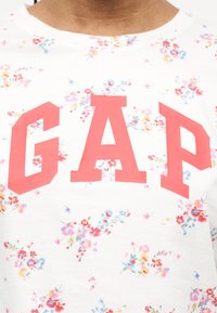 Λευκό πουκάμισο με μικρό πολύχρωμο ανθικό μοτίβο και μεγάλα έντονα κόκκινα γράμματα που σχηματίζουν το "GAP" στο στήθος.