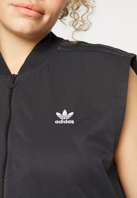 Svart ärmlös jacka i slätt tyg. Har en dragkedja fram och en vit Adidas-logga på vänster bröst.